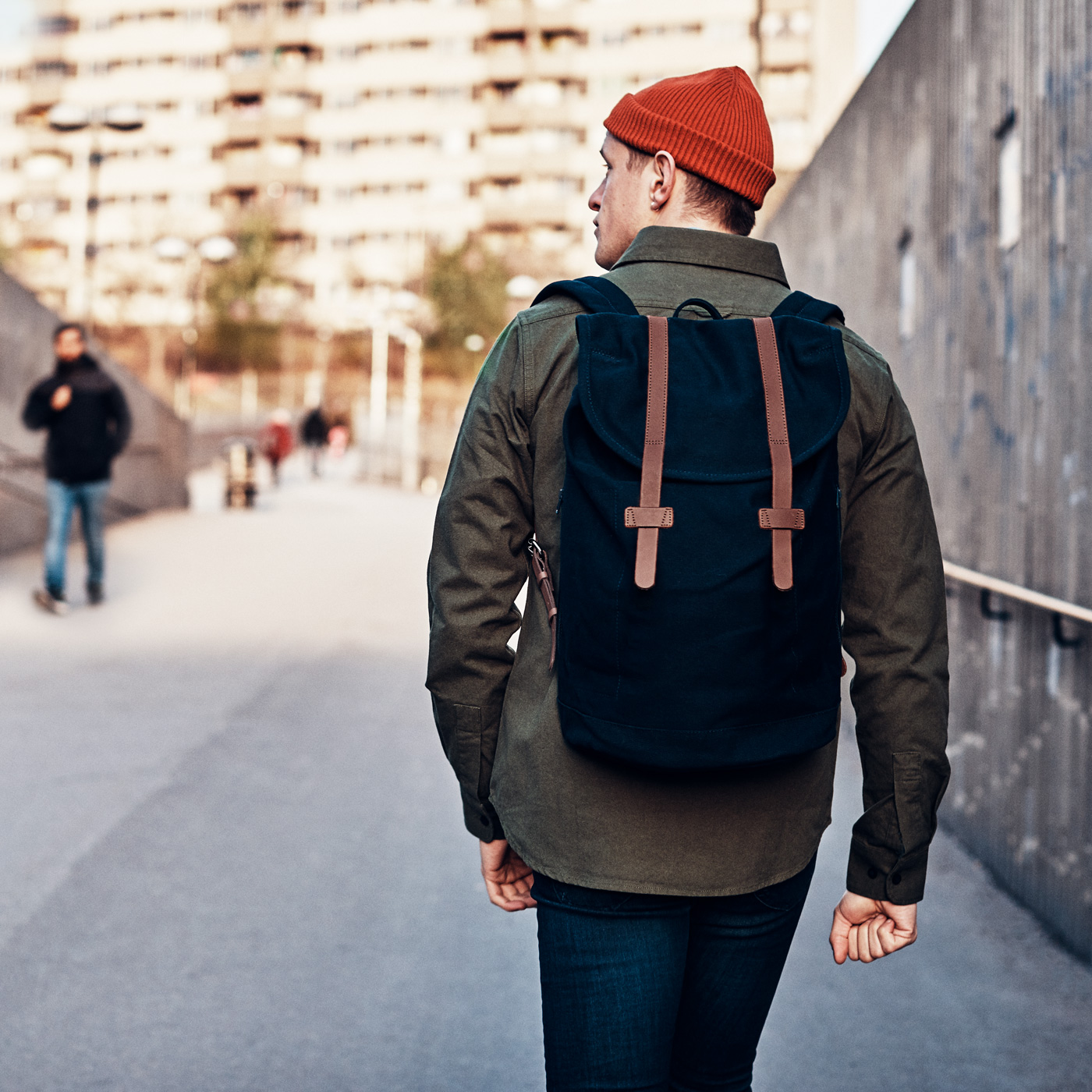En ung man med grön jacka, röd mössa och svart ryggsäck syns bakifrån. Han är på väg hem från tunnelbanan i Bredäng, Stockholm.